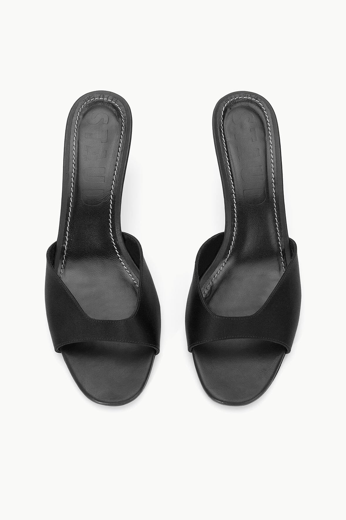 A pair of black Staud Brigitte Mules in satin with open toes and wide straps across the front, featuring a sleek, minimalist design with a slightly higher heel that's perfect for versatile dressing. The insoles have visible stitching around the edges, set against a plain, light-colored background.