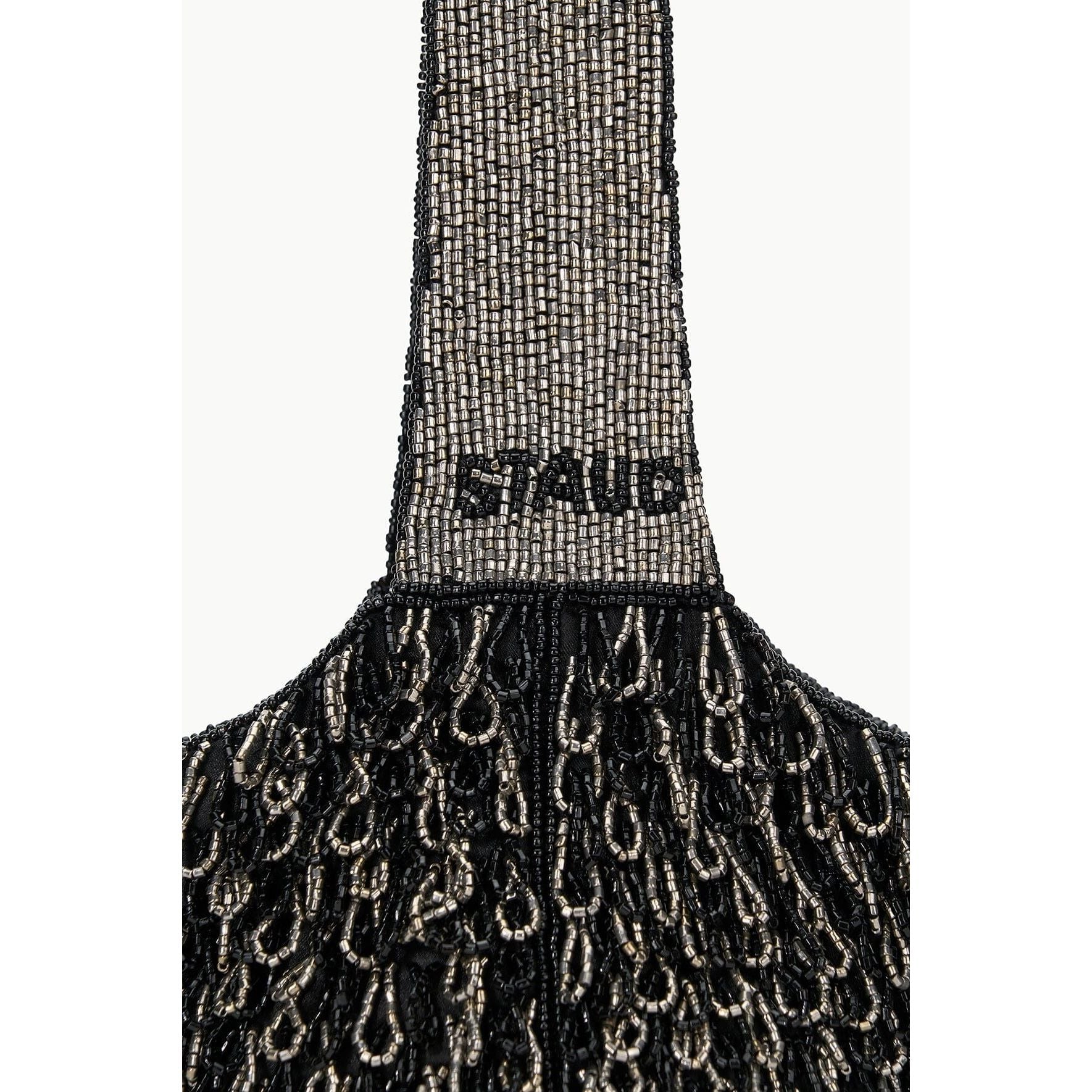 A close-up of the Staud Haruko Beaded Bag showcases its exquisite black and silver beadwork on a textured surface. Crafted from vegan leather, the handle is adorned with densely arranged beads spelling out "STAUD," exuding a luxurious and elegant look.