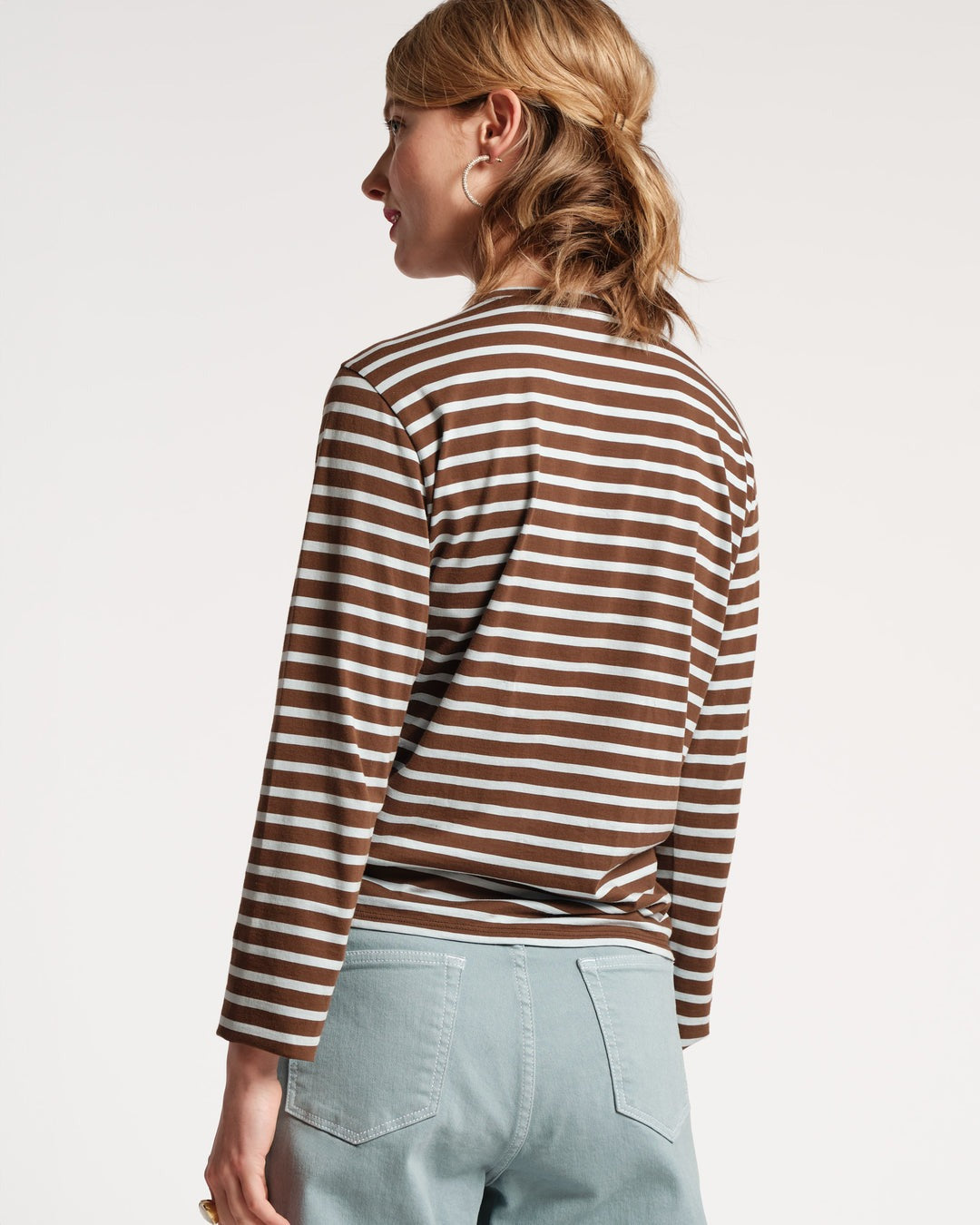 A person with light brown hair is shown from the back, wearing the fitted Frances Valentine Striped Top made of 100% Pima Cotton in Chocolate Light Blue and a pair of light blue jeans. They have a large hoop earring in one ear and are holding something small in one hand, perfect for an autumn wardrobe.