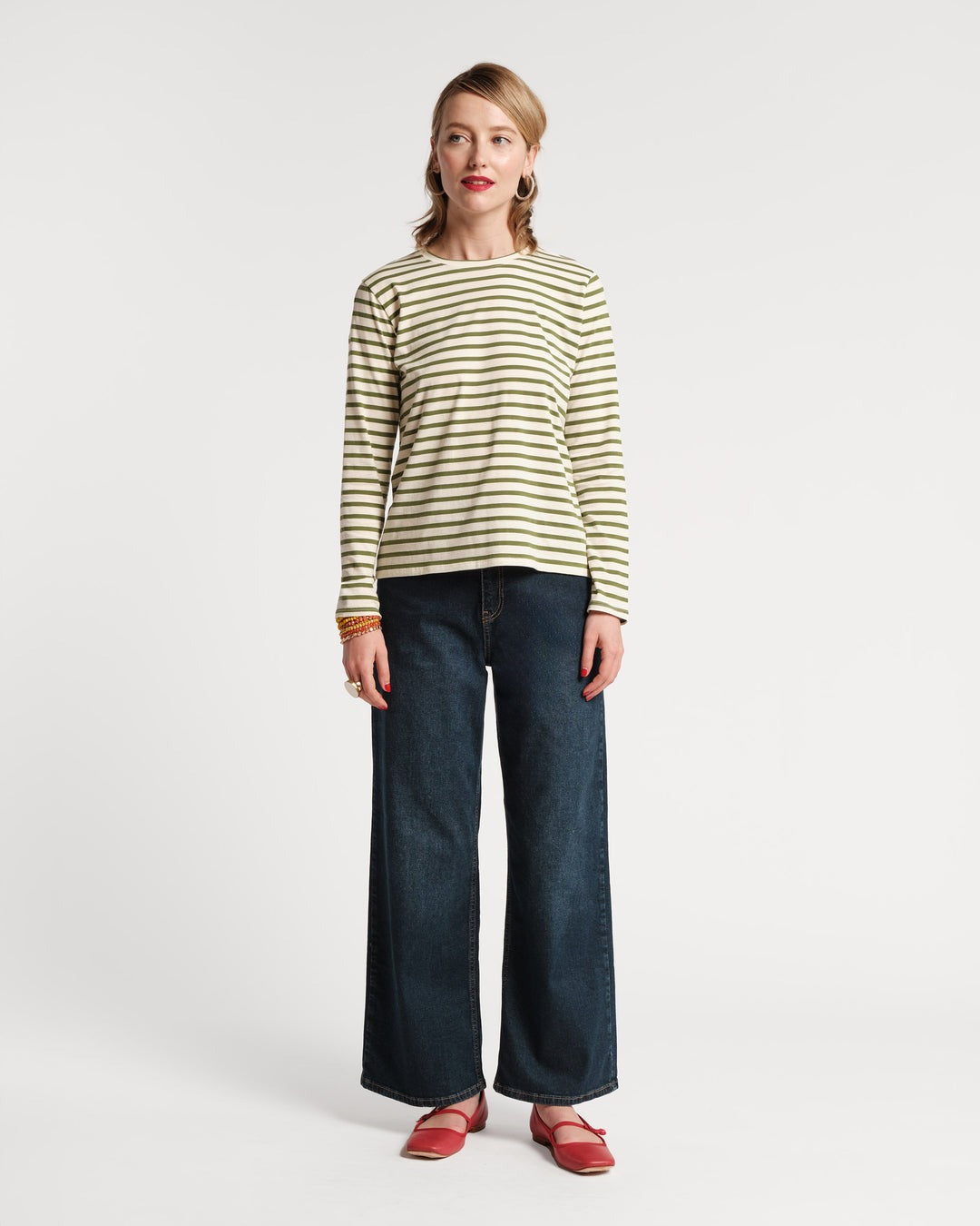 A woman stands against a plain white background, wearing the Frances Valentine Long Sleeve Striped Tee Shirt in Oyster Green paired with wide-leg jeans and red shoes. Her shoulder-length hair frames her face as she smiles slightly, accessorized with bracelets. Perfect for an autumn wardrobe, her outfit creates an elegant elongated silhouette.