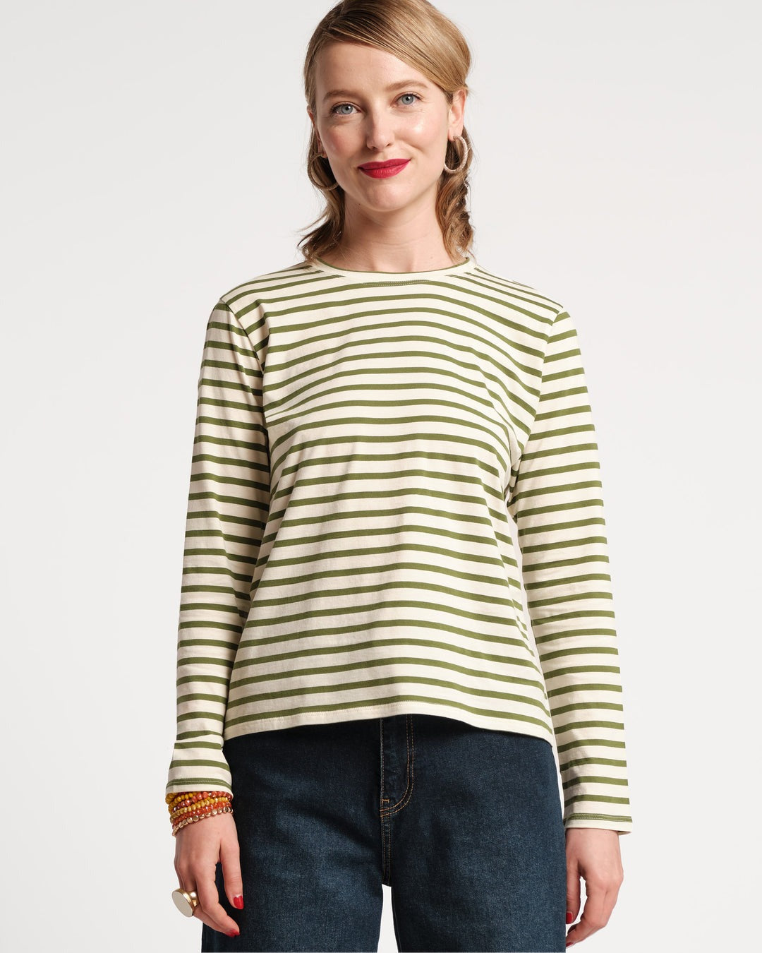 A woman with shoulder-length blonde hair stands against a plain background, smiling slightly. She is wearing the Frances Valentine Long Sleeve Striped Tee Shirt in Oyster Green, dark jeans, a gold ring, gold hoop earrings, and red lipstick—a chic addition to any autumn wardrobe.