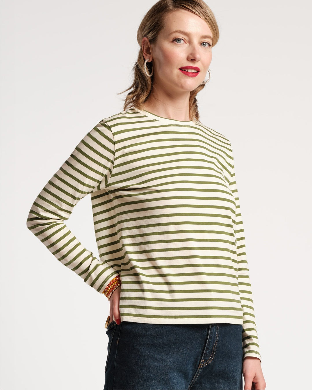 A person with shoulder-length hair stands confidently with one hand on their hip. They wear the Frances Valentine Long Sleeve Striped Tee Shirt Oyster Green and dark jeans, creating an elongated silhouette perfect for an autumn wardrobe. Hoop earrings frame their smiling face, with red lipstick adding a pop of color against the plain white background.