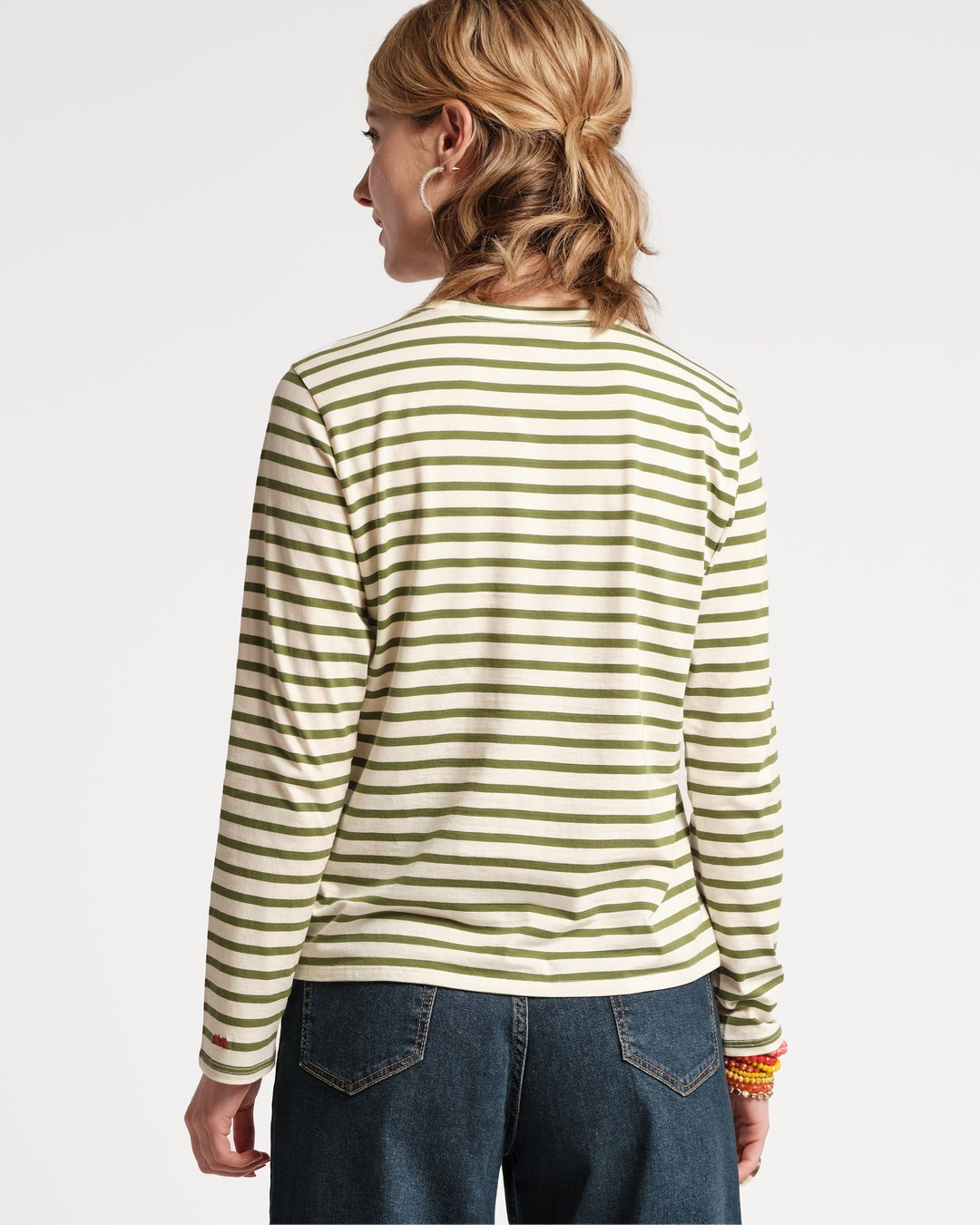 A person with blond hair, styled in loose waves, is standing facing away from the camera, wearing a Frances Valentine Long Sleeve Striped Tee Shirt Oyster Green from Frances Valentine paired with dark jeans. They have several colorful bracelets on their right wrist. The background is plain and light-colored.