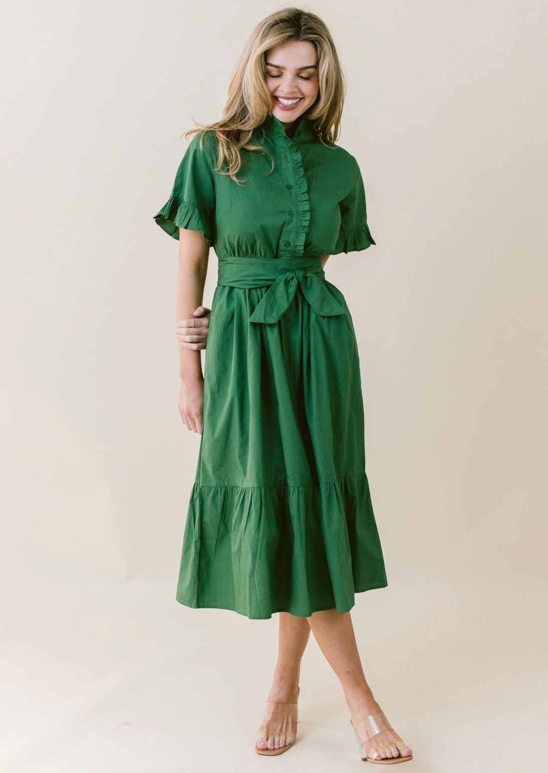 A woman with long hair is smiling and posing in the LaRoque Anna Dress, a green, short-sleeved, button-up, knee-length dress featuring a waistband tie and a ruffled hem. The poly georgette fabric adds a delicate touch. She is standing in front of a neutral background and wearing beige sandals.