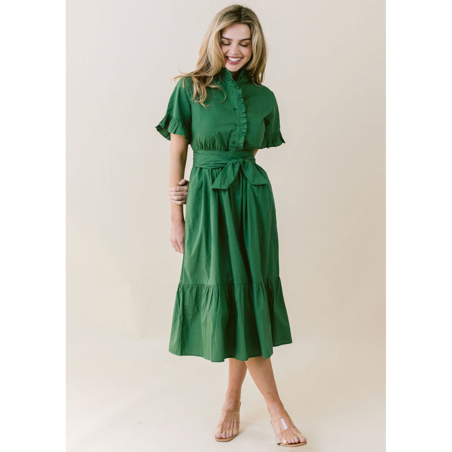 A woman with long hair is smiling and posing in the LaRoque Anna Dress, a green, short-sleeved, button-up, knee-length dress featuring a waistband tie and a ruffled hem. The poly georgette fabric adds a delicate touch. She is standing in front of a neutral background and wearing beige sandals.