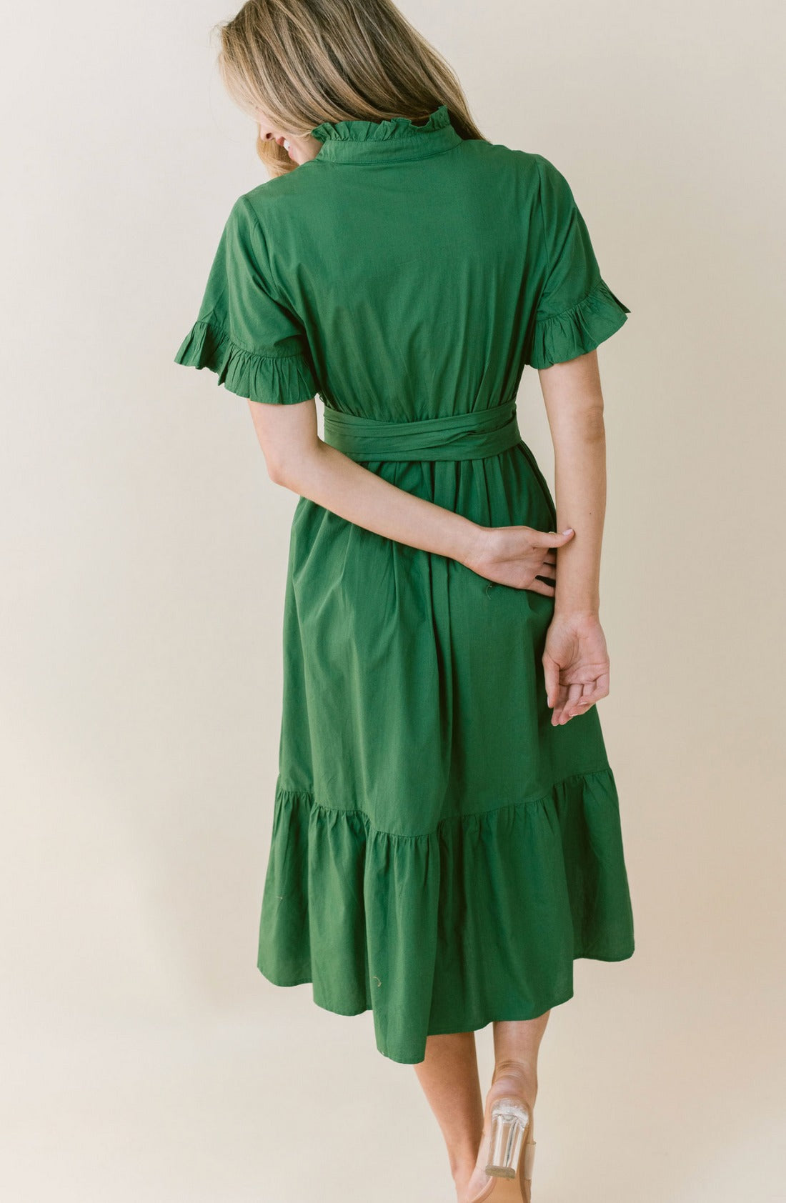 A woman with blond hair, seen from behind, is wearing a green, knee-length LaRoque Anna Dress made of elegant poly georgette. The dress features elastic puff sleeves and a ruffled hem. A belt is tied at the waist. She stands on one leg, with the other heel raised slightly, her arm held behind her back.