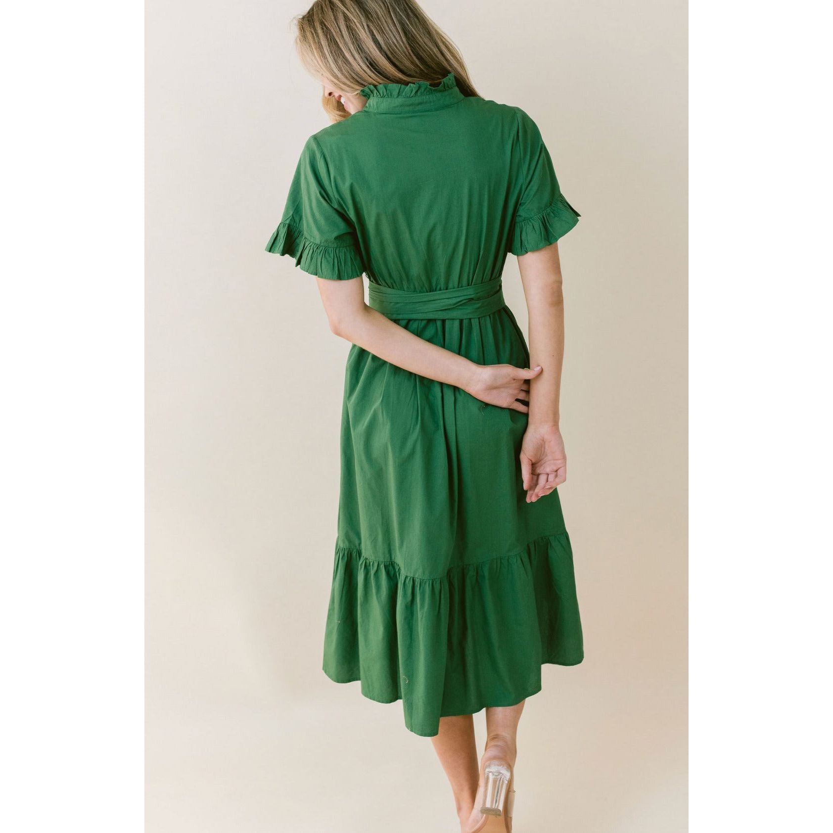 A woman with blond hair, seen from behind, is wearing a green, knee-length LaRoque Anna Dress made of elegant poly georgette. The dress features elastic puff sleeves and a ruffled hem. A belt is tied at the waist. She stands on one leg, with the other heel raised slightly, her arm held behind her back.