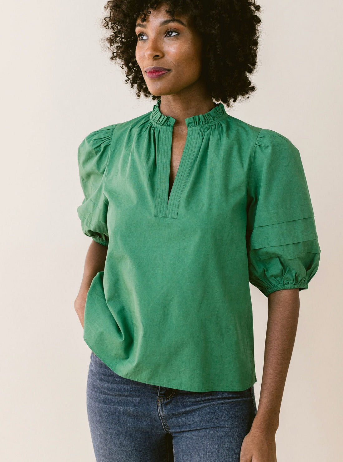 A woman with curly hair is wearing the LaRoque Alix Top Green, which features puffed voile sleeves and a high collar with a V-shaped notch. She has one hand in the pocket of her blue jeans and is standing against a plain beige background.