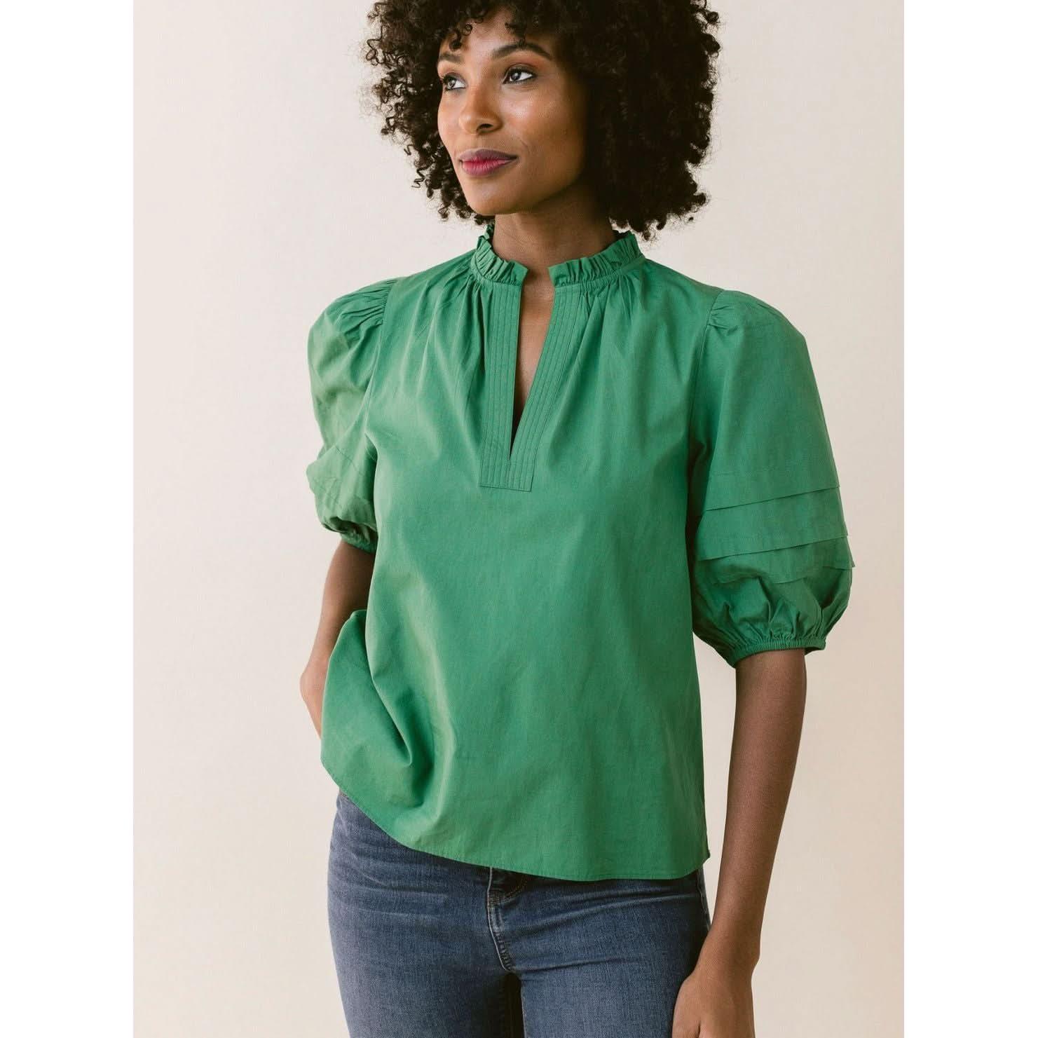 A woman with curly hair is wearing the LaRoque Alix Top Green, which features puffed voile sleeves and a high collar with a V-shaped notch. She has one hand in the pocket of her blue jeans and is standing against a plain beige background.