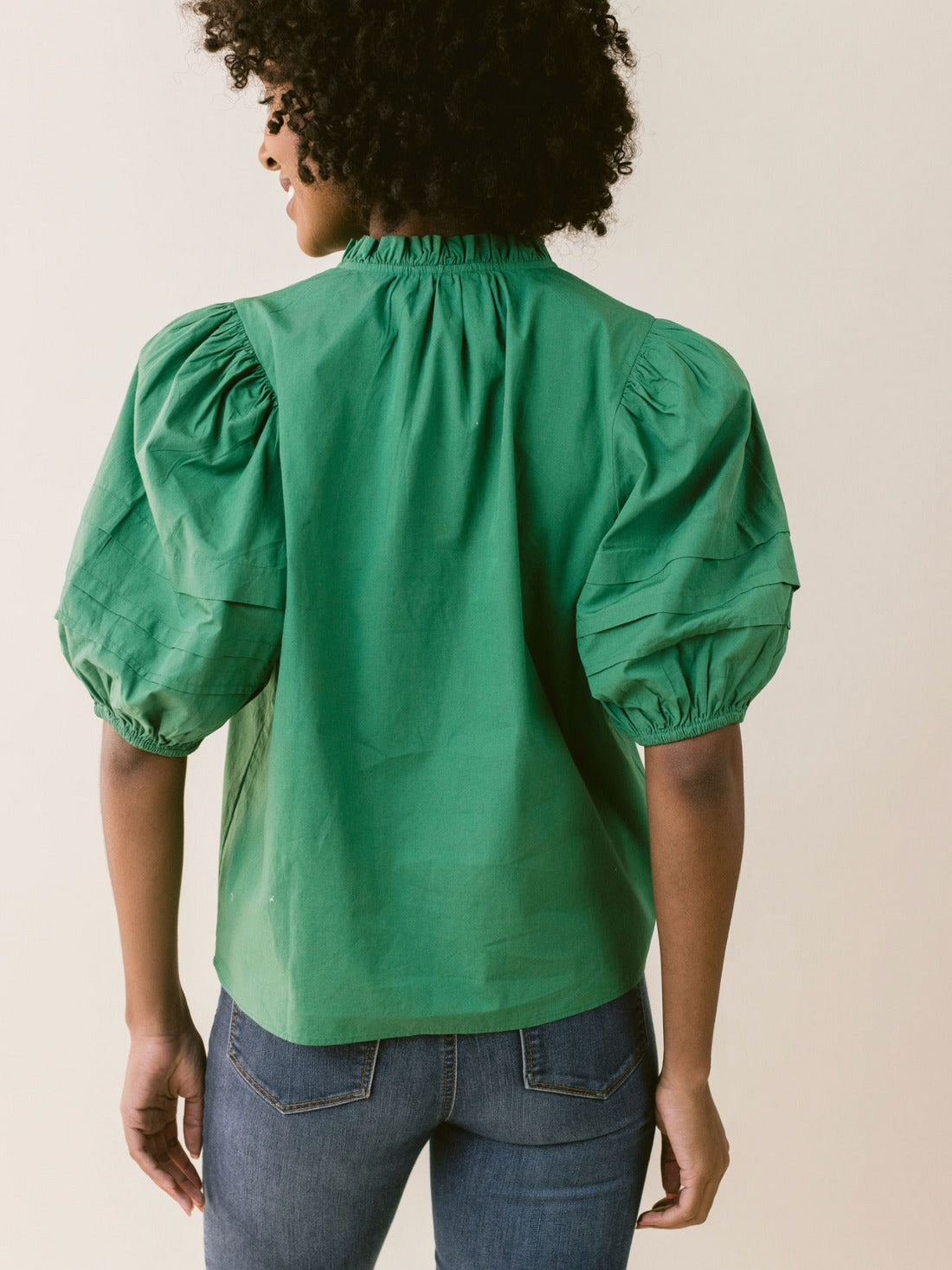 A person with curly hair is standing with their back to the camera. They are wearing the LaRoque Alix Top Green, a bright green blouse from LaRoque featuring puffed short sleeves and elastic cuffs, paired with blue denim jeans. The top has a gathered neckline and a loose fit. The background is a neutral, off-white color.