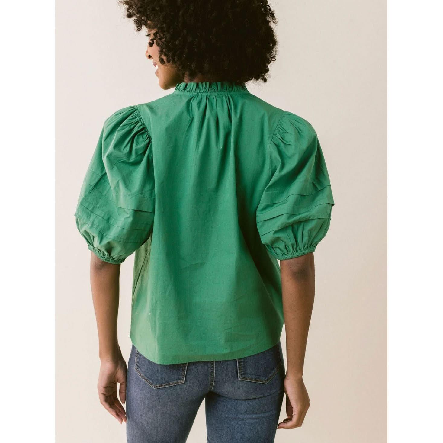 A person with curly hair is standing with their back to the camera. They are wearing the LaRoque Alix Top Green, a bright green blouse from LaRoque featuring puffed short sleeves and elastic cuffs, paired with blue denim jeans. The top has a gathered neckline and a loose fit. The background is a neutral, off-white color.