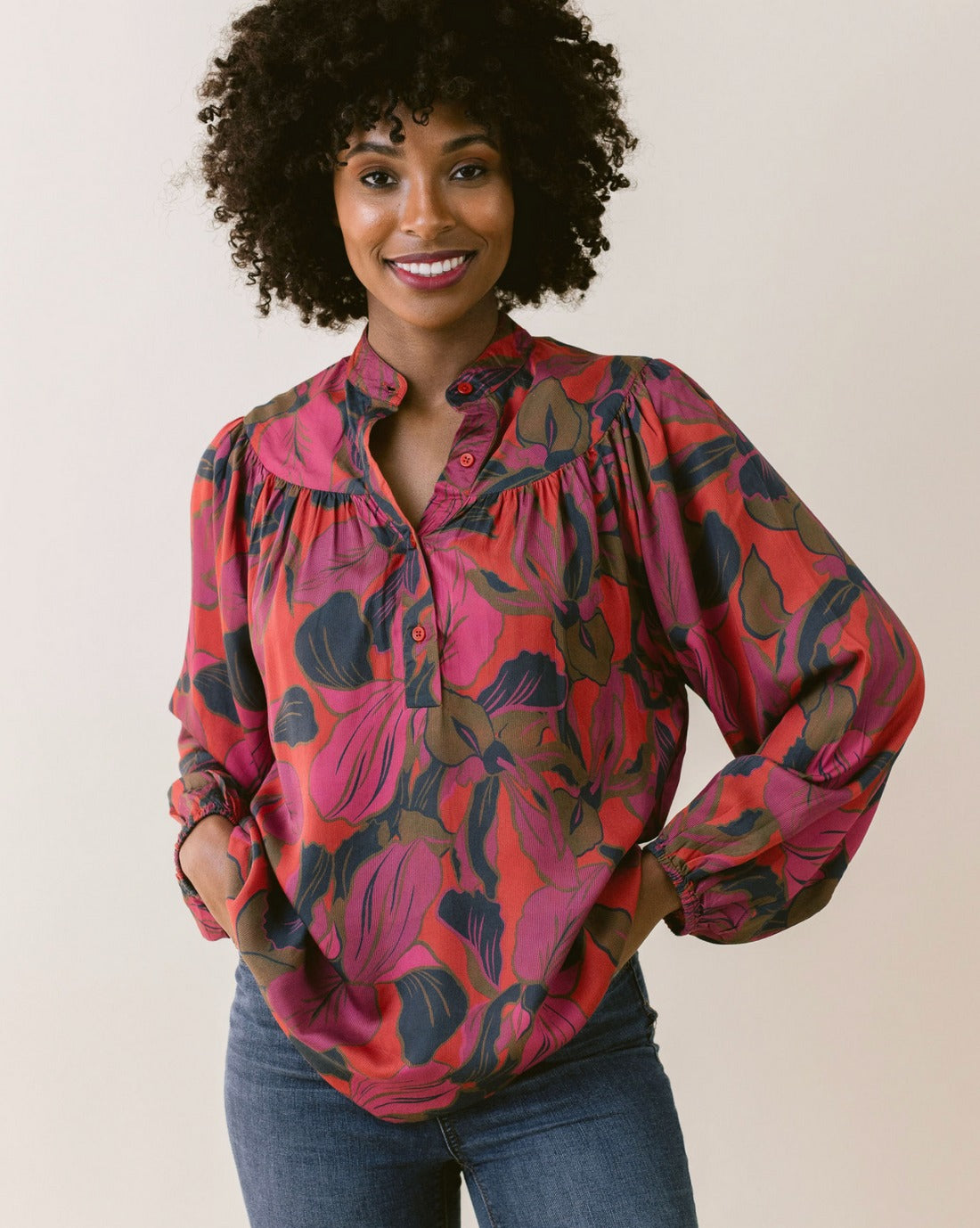 A woman with curly hair smiles at the camera, wearing the LaRoque Bailey Blouse, a colorful floral blouse in red, pink, and green tones. The blouse features an open neckline and elastic cuffs, paired elegantly with blue jeans. She stands in front of a plain background with her hands resting on her hips.