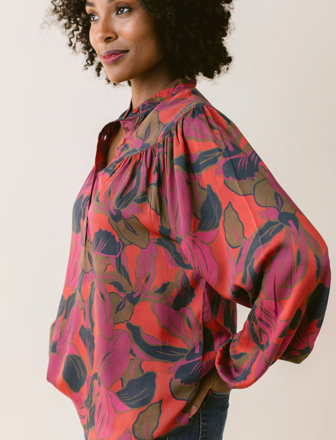 A woman with curly hair is smiling and posing sideways. She is wearing the LaRoque Bailey Blouse, a vibrant, long-sleeved top with an open neckline and a bold, colorful floral pattern in shades of red, pink, and purple. The elastic cuffs add a touch of elegance. The background is a plain, light color.