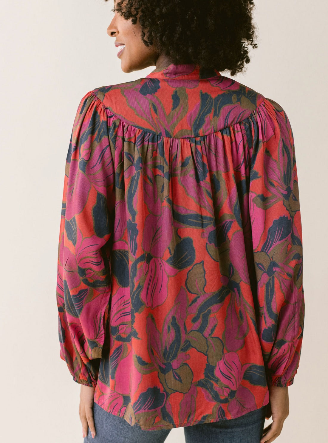 A person is shown from the back wearing the LaRoque Bailey Blouse, made of Desert Palm cotton with a vibrant floral pattern in shades of red, pink, and dark blue. The blouse features an open neckline, long billowy sleeves with elastic cuffs, and a loose fit. The person has curly hair and is smiling against a light-colored background.