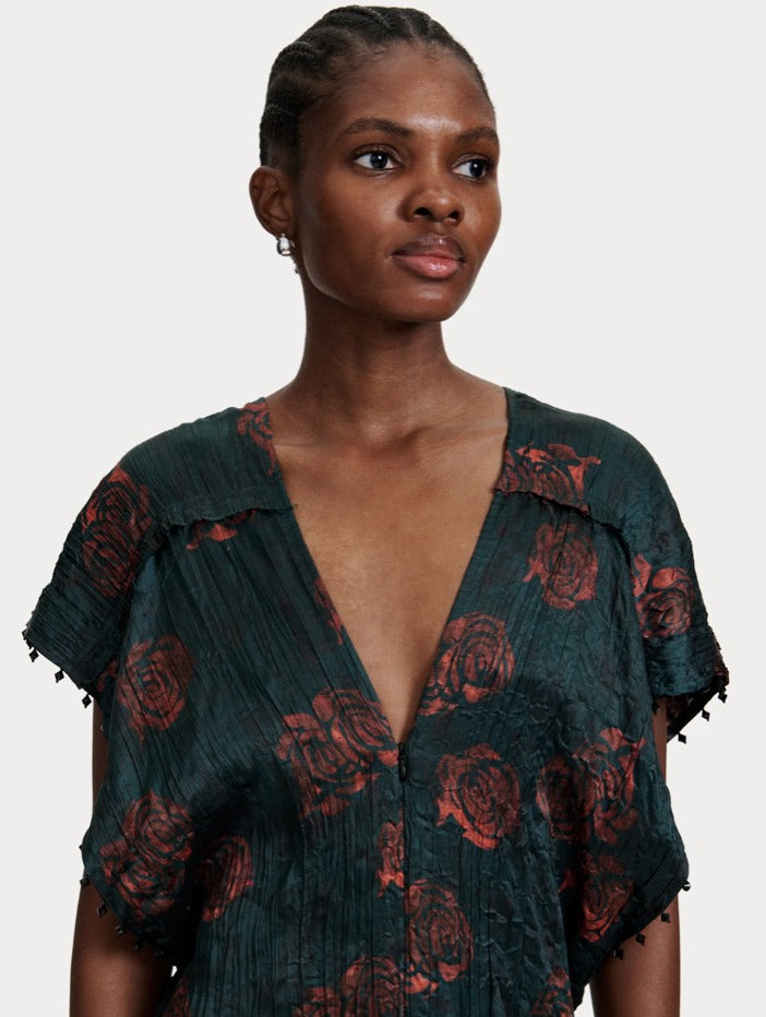 A woman with short, braided hair is wearing the Rachel Comey Sisu Dress from Rachel Comey, a V-neck, short-sleeve piece in dark green adorned with a red floral pattern. She stands against a plain off-white background, exuding effortless elegance reminiscent of a tunic-style dress.