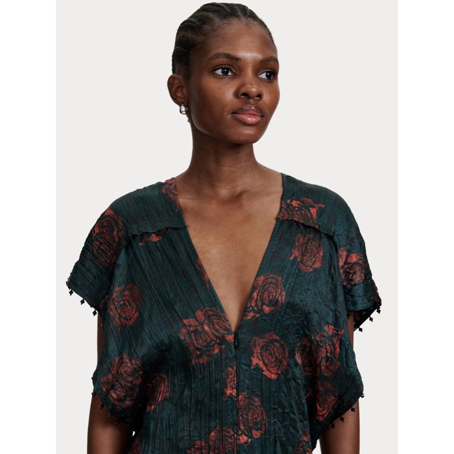 A woman with short, braided hair is wearing the Rachel Comey Sisu Dress from Rachel Comey, a V-neck, short-sleeve piece in dark green adorned with a red floral pattern. She stands against a plain off-white background, exuding effortless elegance reminiscent of a tunic-style dress.