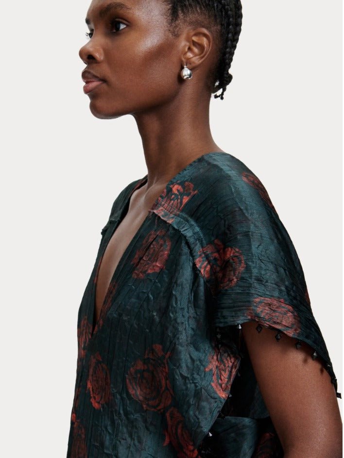 A side profile of a person with short, braided hair wearing pearl earrings and the Rachel Comey Sisu Dress. The dark, crinkled viscose dress features rose designs, a deep V-neck, and jagged sleeves adorned with small beads. The background is plain light grey.