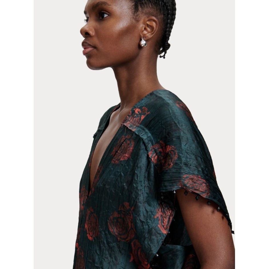 A side profile of a person with short, braided hair wearing pearl earrings and the Rachel Comey Sisu Dress. The dark, crinkled viscose dress features rose designs, a deep V-neck, and jagged sleeves adorned with small beads. The background is plain light grey.