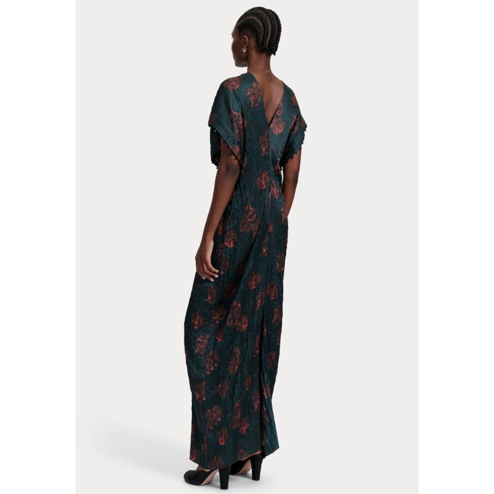 A person with braided hair, seen from behind, is wearing the Rachel Comey Sisu Dress, a dark green crinkled viscose dress with a floral pattern and black heels. The dress features a deep V-back and a flowing, floor-length design.