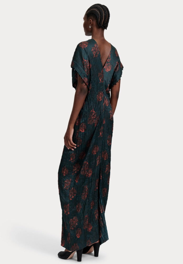 A person with braided hair, seen from behind, is wearing the Rachel Comey Sisu Dress, a dark green crinkled viscose dress with a floral pattern and black heels. The dress features a deep V-back and a flowing, floor-length design.