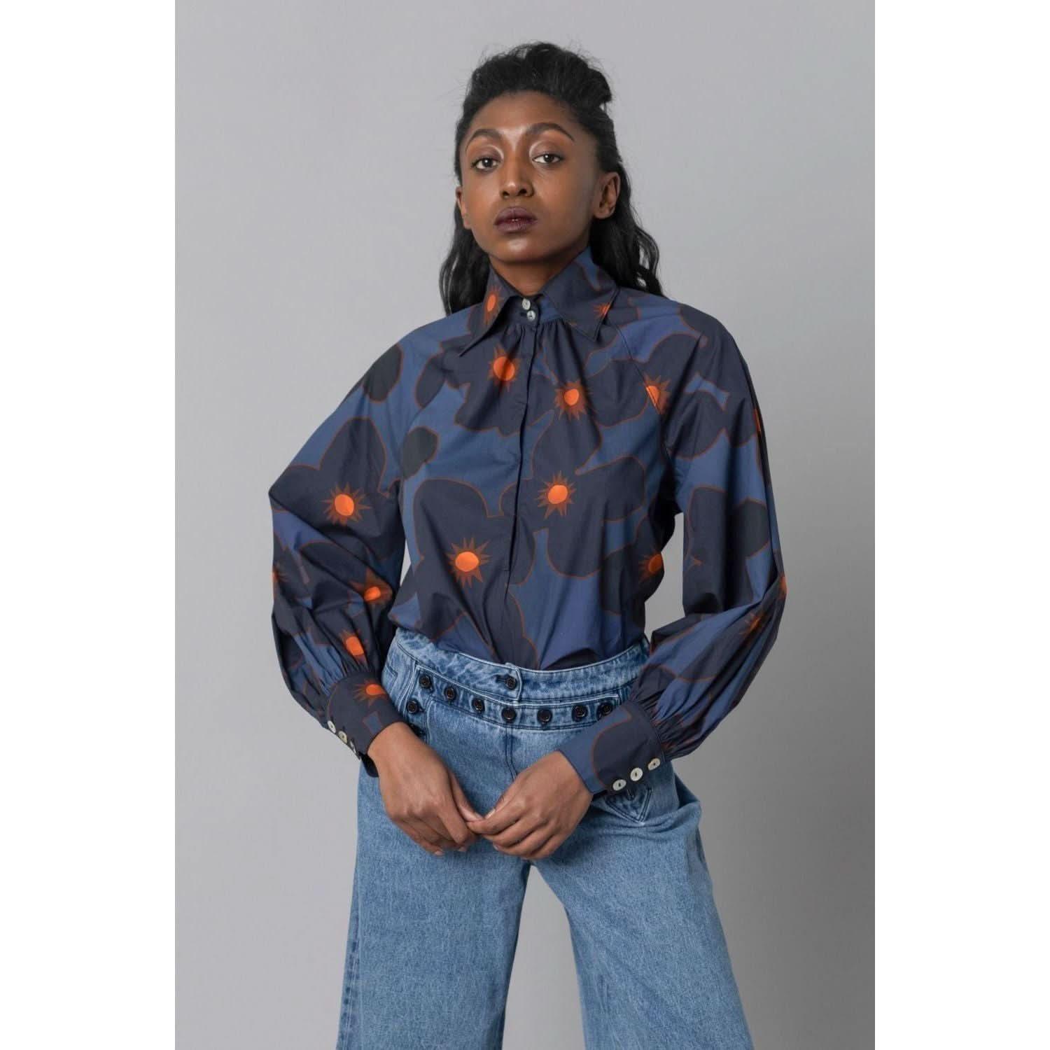 A person with long dark hair stands confidently against a gray background, wearing the Hope for Flowers Pull Over Shirt Kiki Floral by Hope for Flowers by Tracy Reece. The blouse features a vibrant Kiki Floral design and is made from organic cotton, paired with high-waisted blue jeans. Their hands rest at the waist, reflecting an appreciation for ethical manufacturing practices.