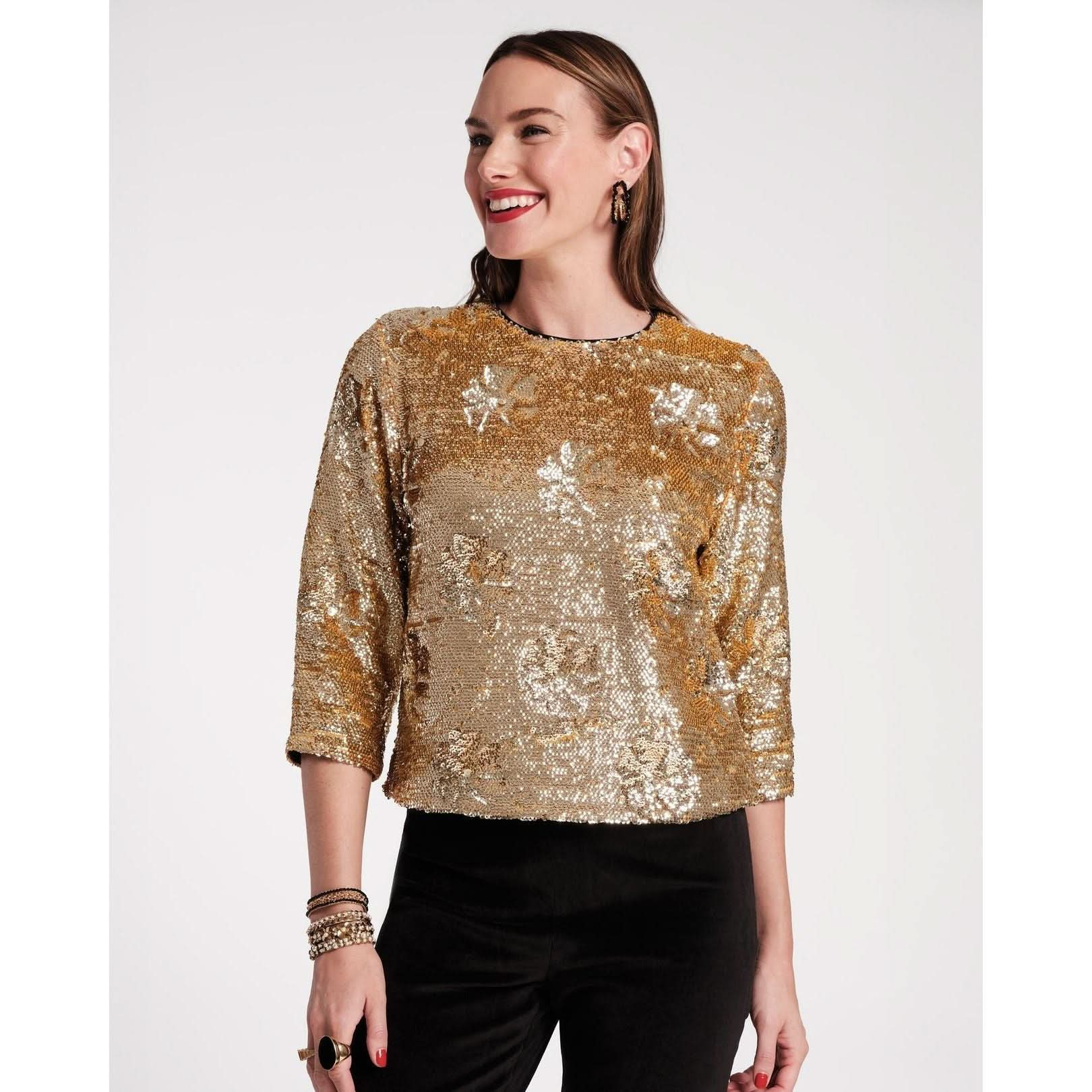 A person smiling, wearing a gold Frances Valentine Taylor Sequin Top Floral with three-quarter sleeves and black pants. The person has long hair and is accessorized with hoop earrings and bracelets. This chic evening ensemble is set against a plain, light-colored background.