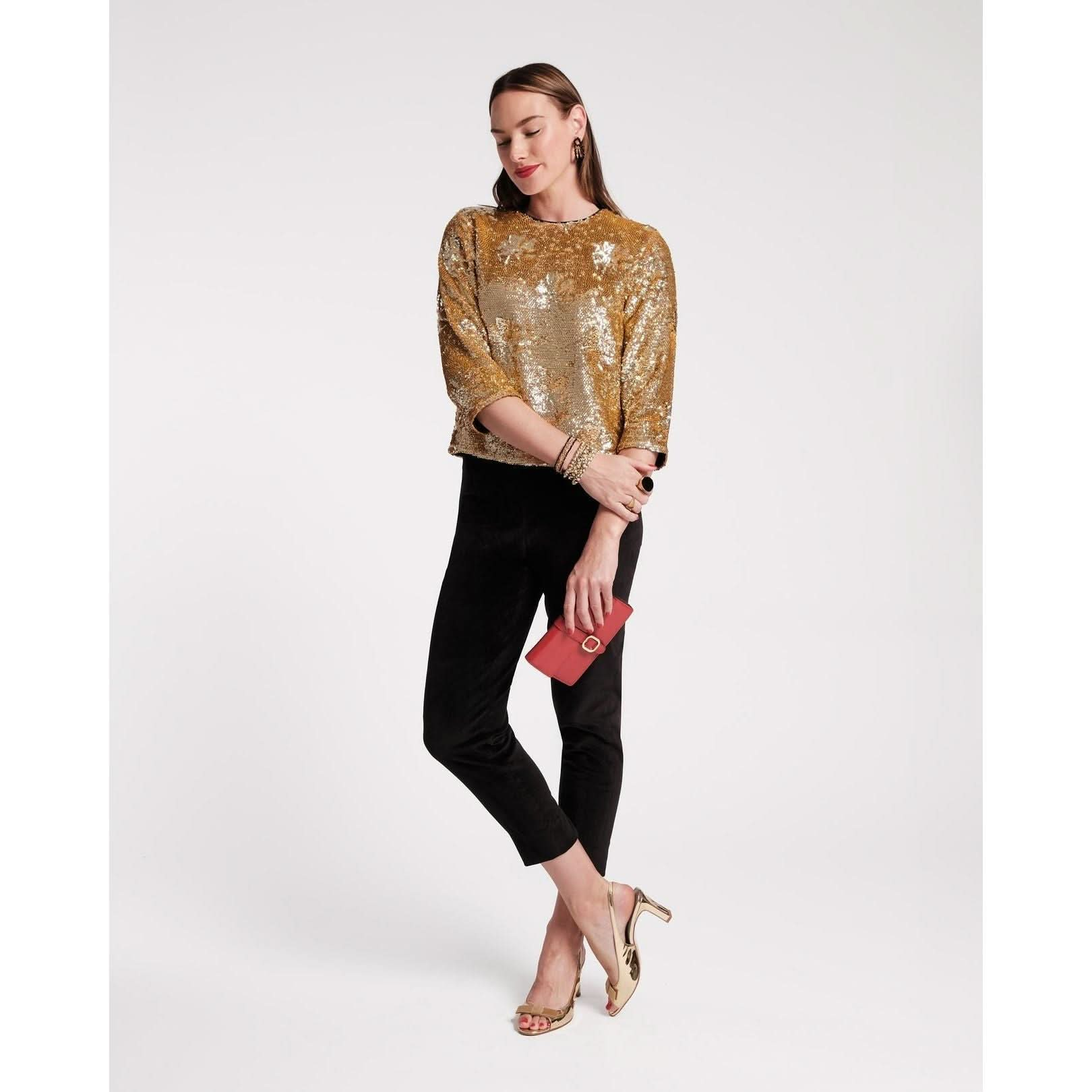 A person stands confidently in gold high heels, black pants, and the Frances Valentine Taylor Sequin Top Floral. They hold a small red purse, wearing gold bracelets, and smiling with closed eyes against a plain background—creating a perfect evening ensemble.