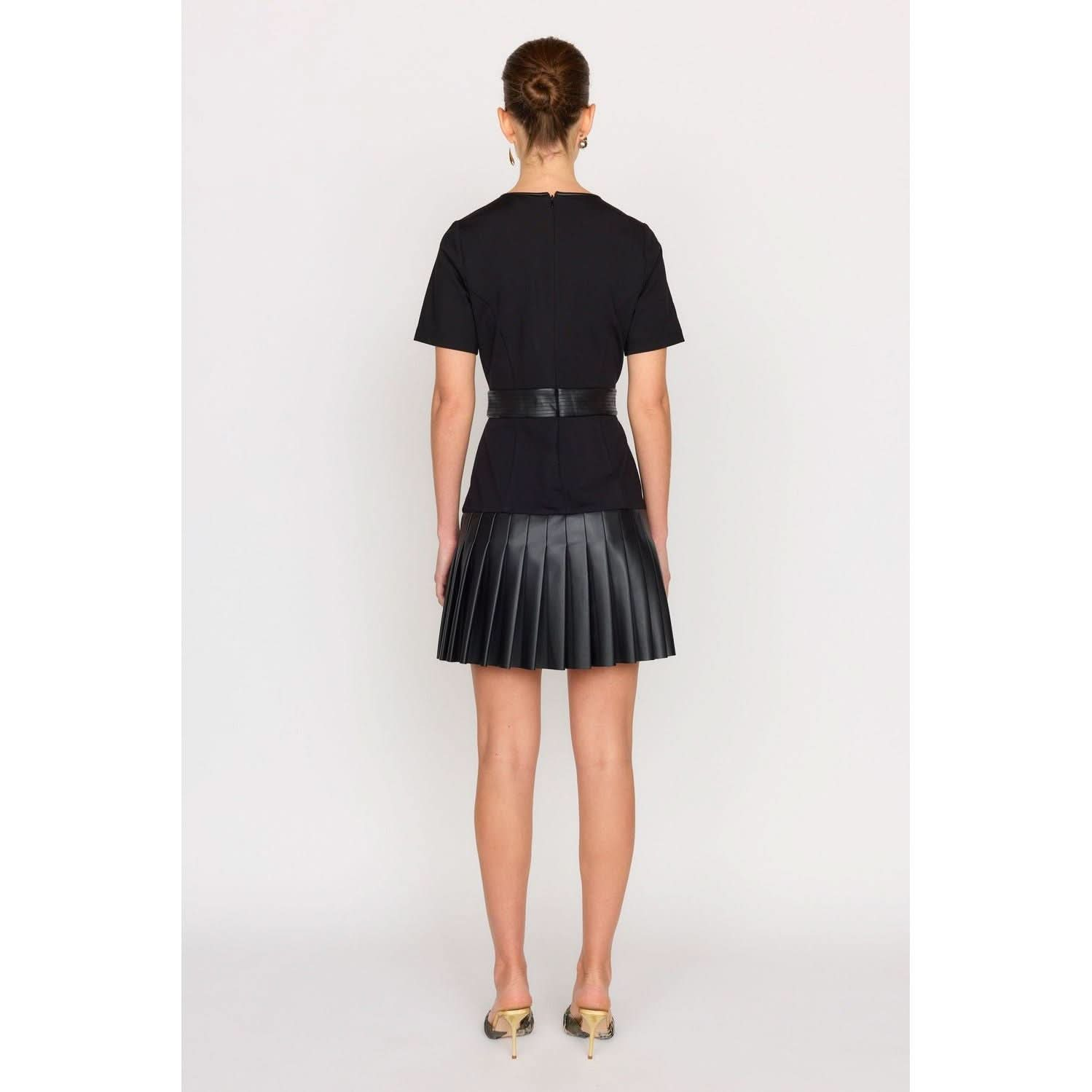 Rear view of a person with tied-up hair wearing a black Christy Lynn Steph Dress, featuring semi-puff sleeves and a pleated skirt, paired with strappy heels. They stand against a plain light background, embodying elegance and sophistication.