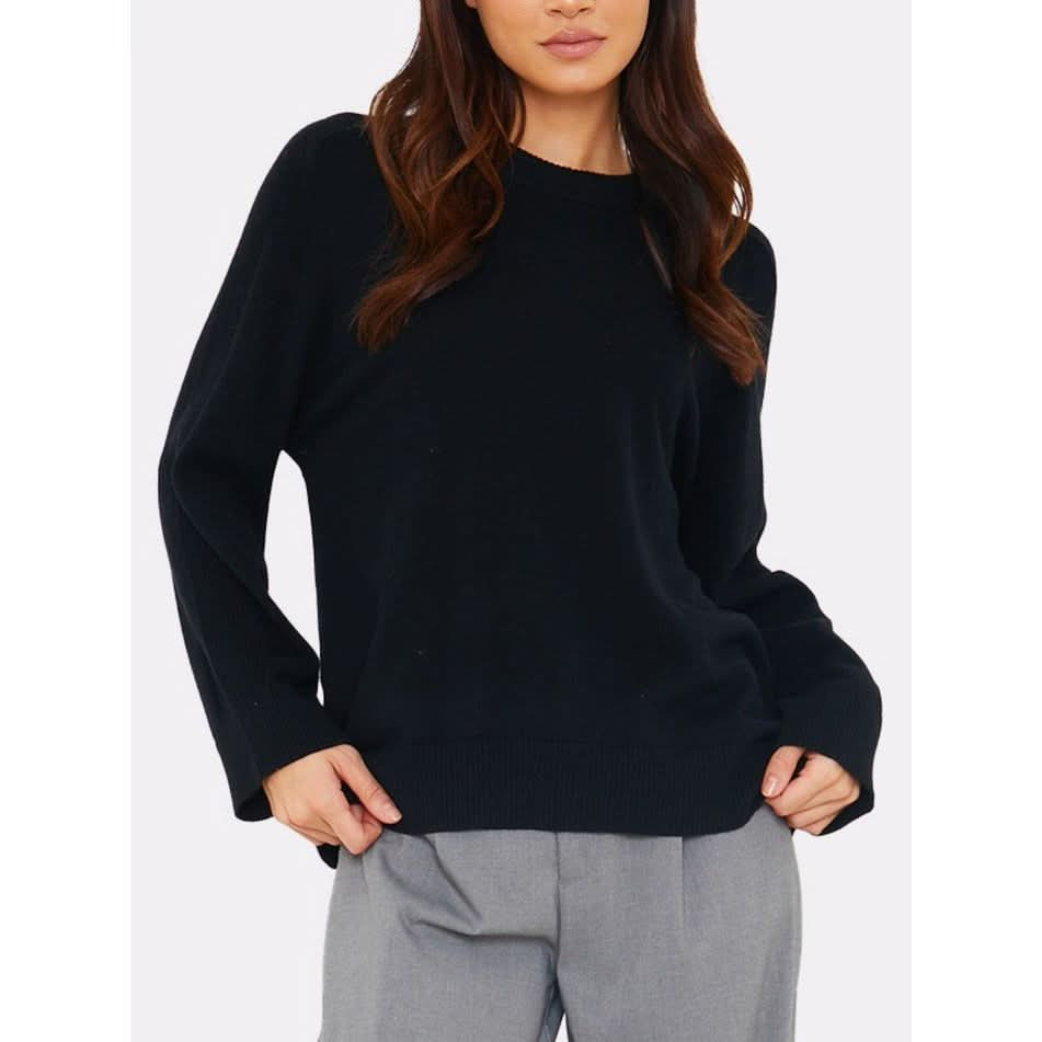 A person wearing a black Brodie Cashmere Saddle Shoulder Rib Knit sweater and gray pants stands against a plain background. The loose-fit sweater from Brodie Cashmere features a crew neckline and long sleeves, with the person's hands casually tucked into their pockets. Their hair is styled in loose waves.