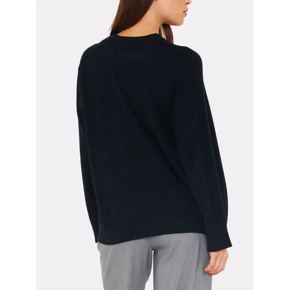 A person wearing the Brodie Cashmere Saddle Shoulder Rib Knit in black with a long sleeve design and a crew neckline, paired with gray pants from the Cotton-Silk Collection, standing and looking to the side against a plain background.