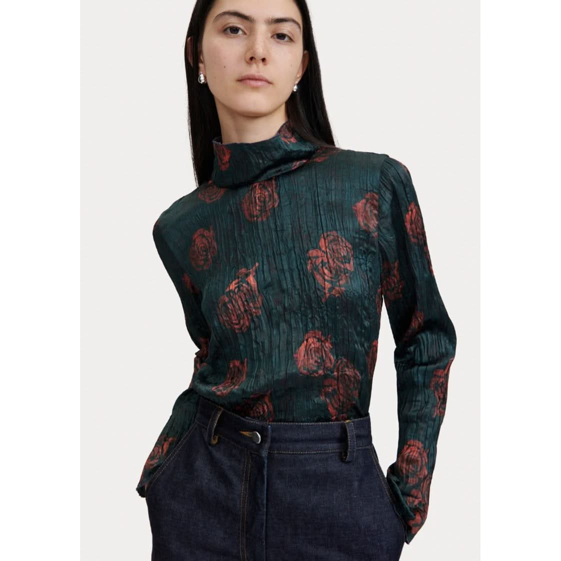 A person stands with hands in pockets, wearing a dark green, long-sleeved Rachel Comey Varda Top adorned with a red rose pattern. They pair it with dark denim jeans. The background is minimal and light-colored.