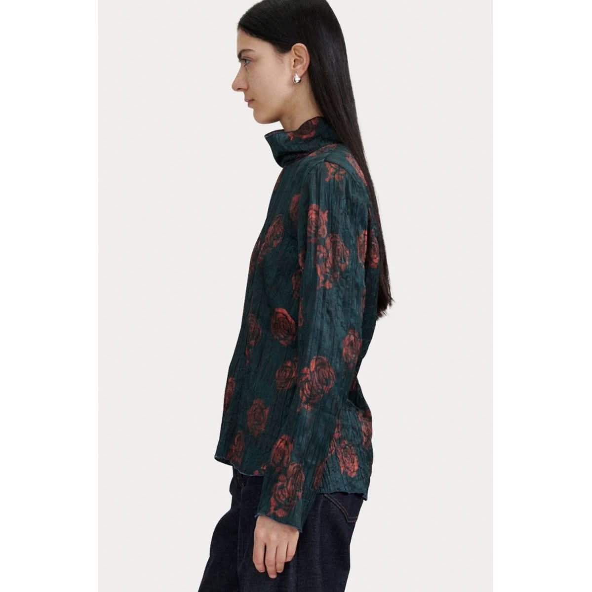 A person with long black hair is standing in a side profile, wearing the Rachel Comey Varda Top, a dark green shirt adorned with rose prints, featuring long sleeves and a mock neck. They are paired with dark pants against a plain, light-colored background.