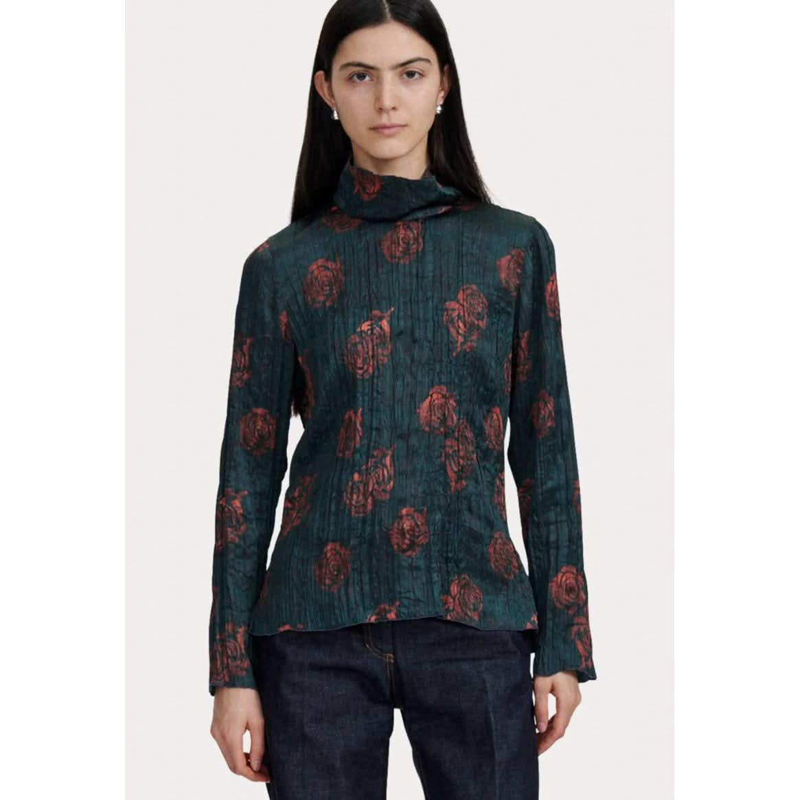 A person with long dark hair is wearing a Rachel Comey Varda Top, a dark green textured mock neck piece adorned with a red floral pattern from the brand Rachel Comey. They are also wearing dark blue jeans. The background is plain and light-colored.