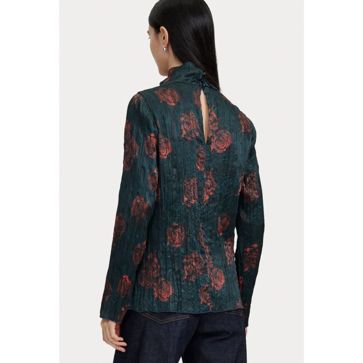 A woman with long, dark hair is shown from the back, wearing the Rachel Comey Varda Top by Rachel Comey. The blouse features a dark green rose print with red floral patterns, a mock neck, and a keyhole opening at the neck secured with buttons. She is also wearing dark pants against a light-colored background.