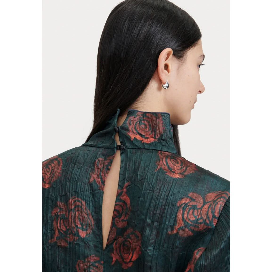 A person with long, dark hair is shown from the back, wearing a Rachel Comey Varda Top. This textured, dark green rose-printed shirt features red floral patterns and boasts a high-neck with button closure and keyhole opening at the back of the neck. The individual also sports small, silver earrings for an elegant evening set.