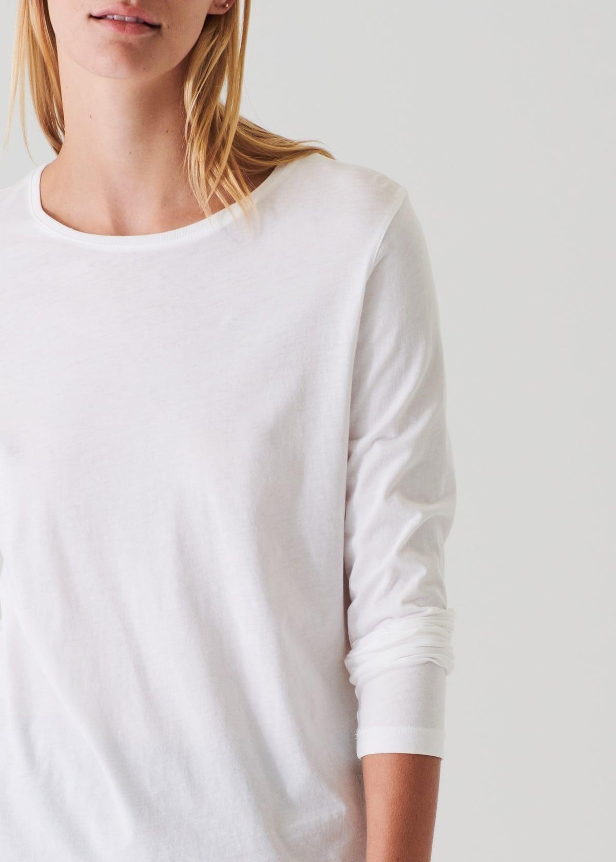 A person with long blonde hair is partially visible from the shoulders up, wearing a Patrick Assaraf Long Sleeve Iconic Boyfriend Crew in white. The classic fit crewneck shirt, made of high-quality Peruvian pima cotton, provides a clean and comfortable appearance. The image is cropped to show only part of their face and upper body, set against a plain background.