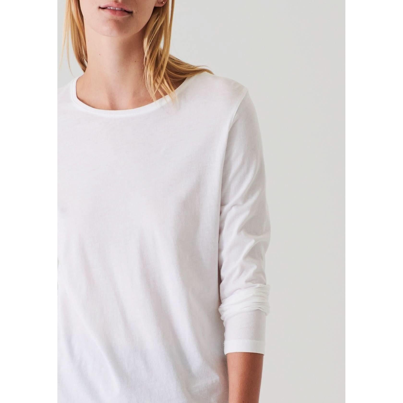 A person with long blonde hair is partially visible from the shoulders up, wearing a Patrick Assaraf Long Sleeve Iconic Boyfriend Crew in white. The classic fit crewneck shirt, made of high-quality Peruvian pima cotton, provides a clean and comfortable appearance. The image is cropped to show only part of their face and upper body, set against a plain background.