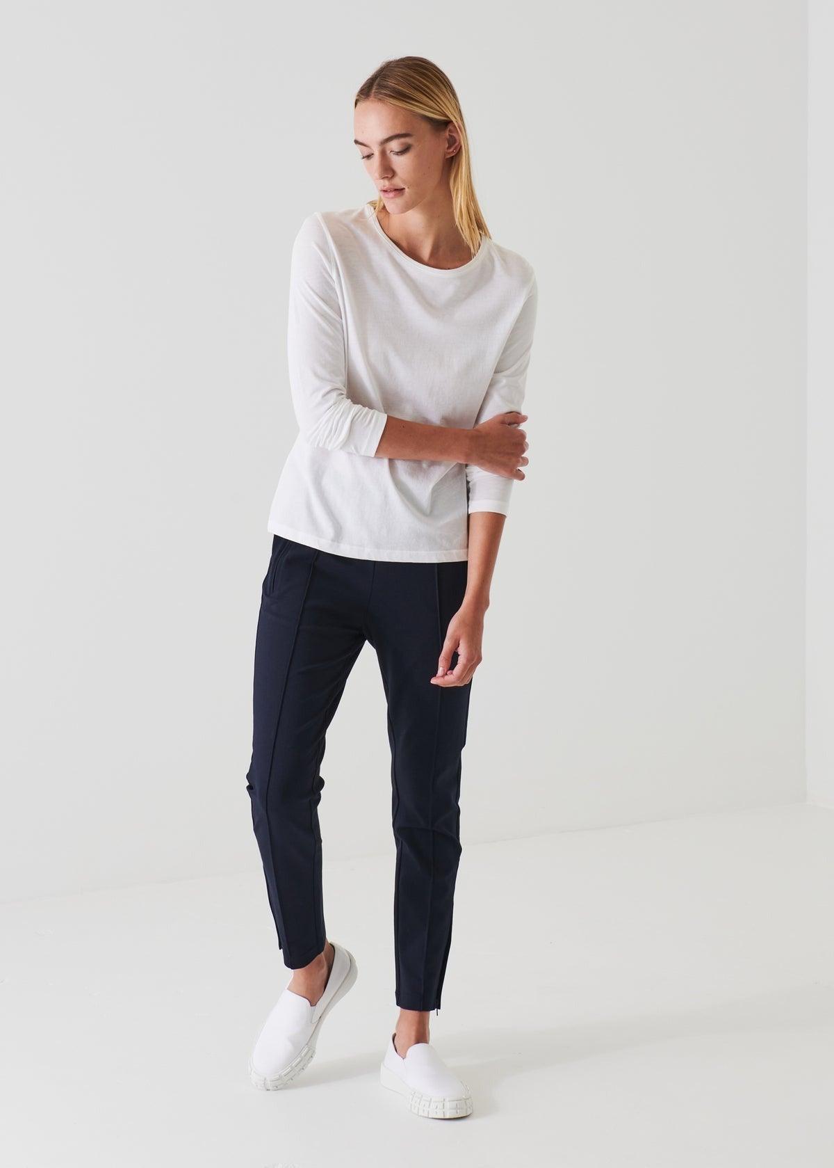 A person with long hair is standing against a plain white background. They are wearing a Patrick Assaraf Long Sleeve Iconic Boyfriend Crew in white, black pants, and white slip-on shoes. Their head is slightly tilted downwards, and their left hand is resting on their right forearm.