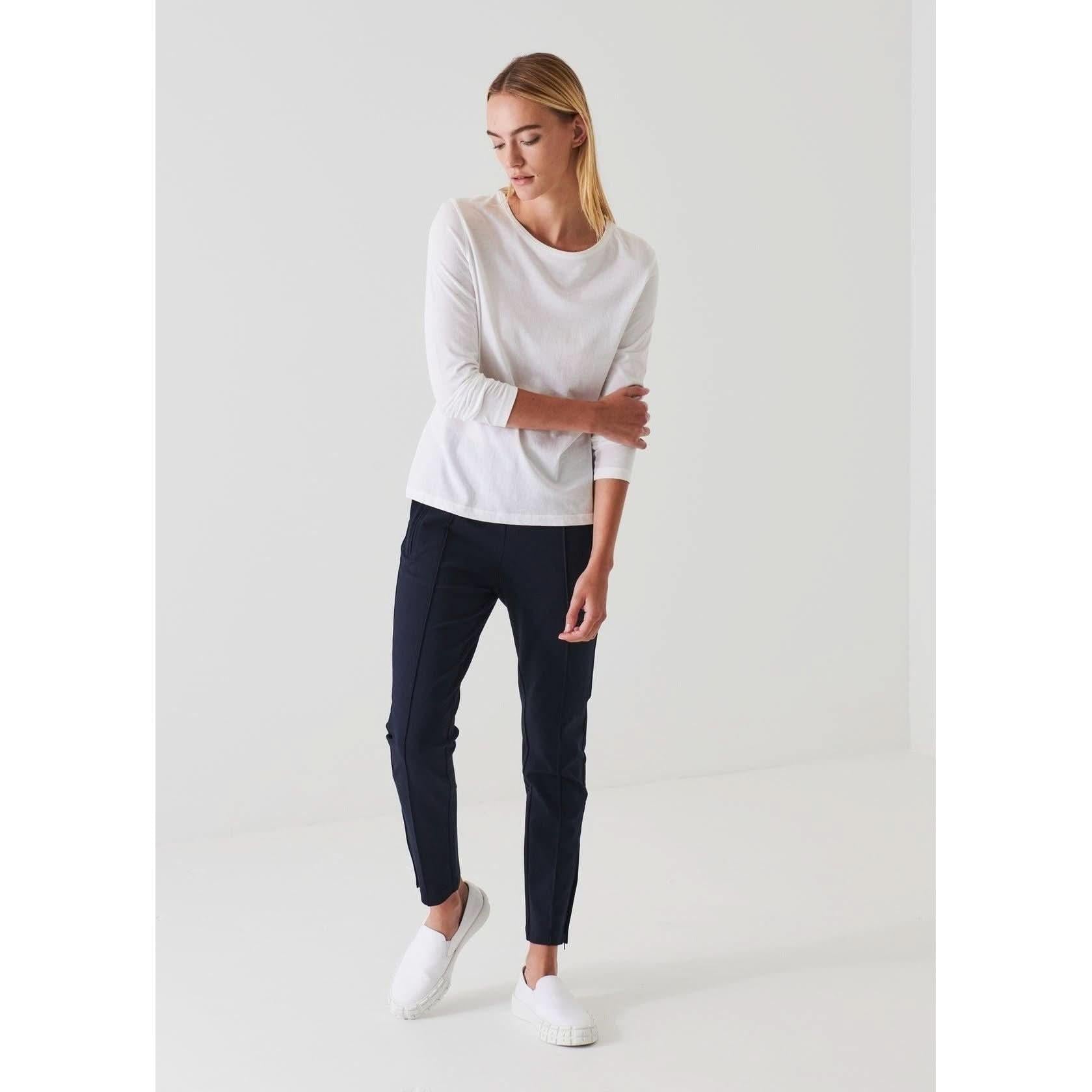 A person with long hair is standing against a plain white background. They are wearing a Patrick Assaraf Long Sleeve Iconic Boyfriend Crew in white, black pants, and white slip-on shoes. Their head is slightly tilted downwards, and their left hand is resting on their right forearm.