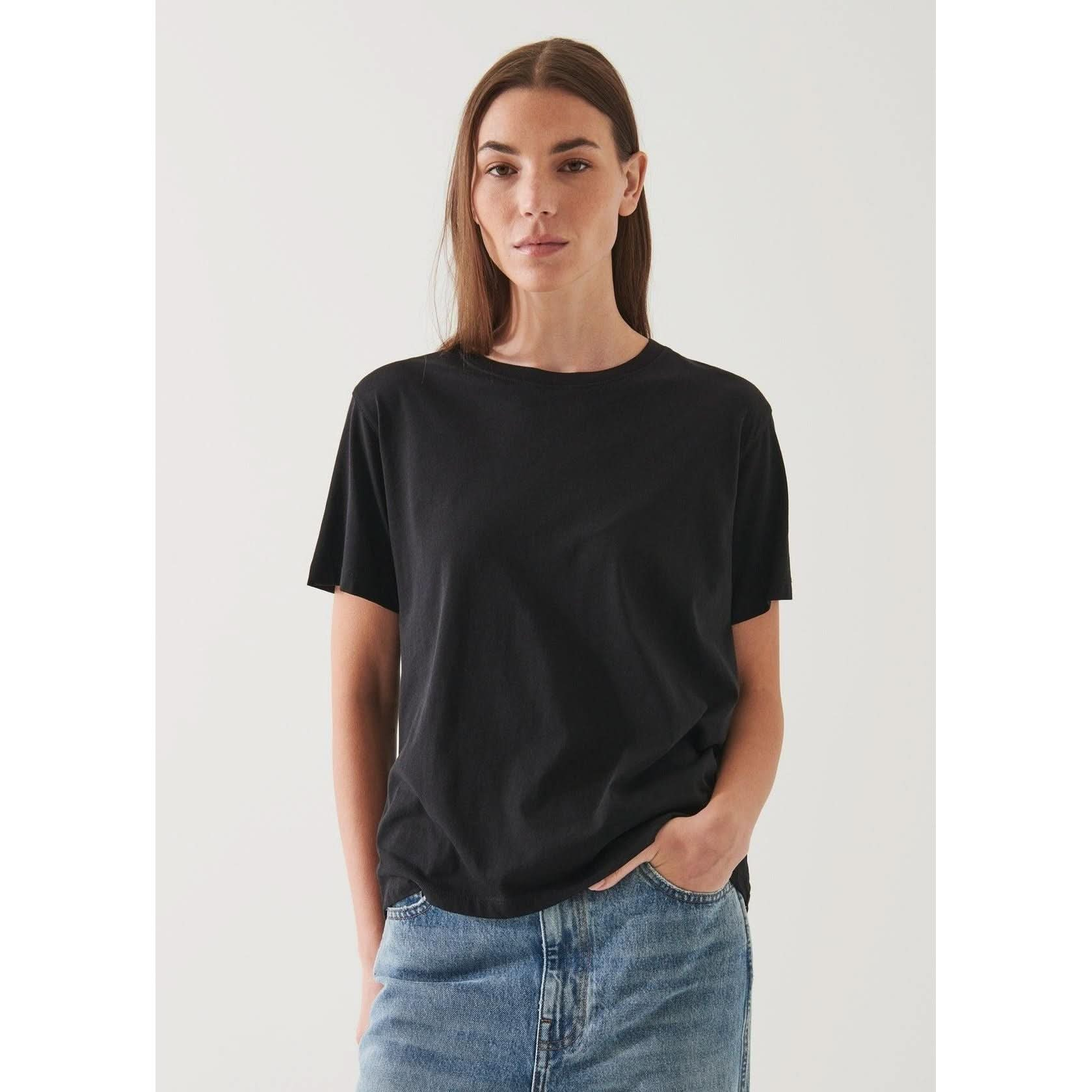 A person with long brown hair wearing an oversized fit, plain black Patrick Assaraf SS Iconic Boyfriend Crew Black T-shirt and light blue jeans stands with one hand in their pocket against a neutral background.