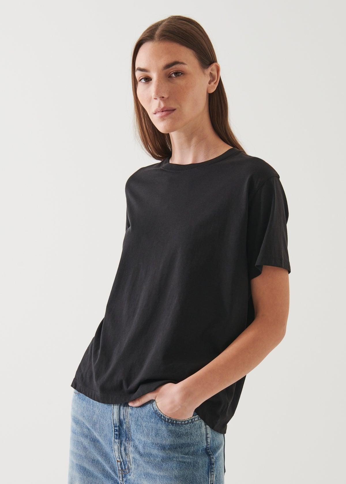 A person with long brown hair, wearing an oversized fit black Patrick Assaraf SS Iconic Boyfriend Crew T-shirt made of soft pima cotton and light blue jeans, stands against a plain light background with one hand in a pocket, looking towards the camera with a neutral expression.