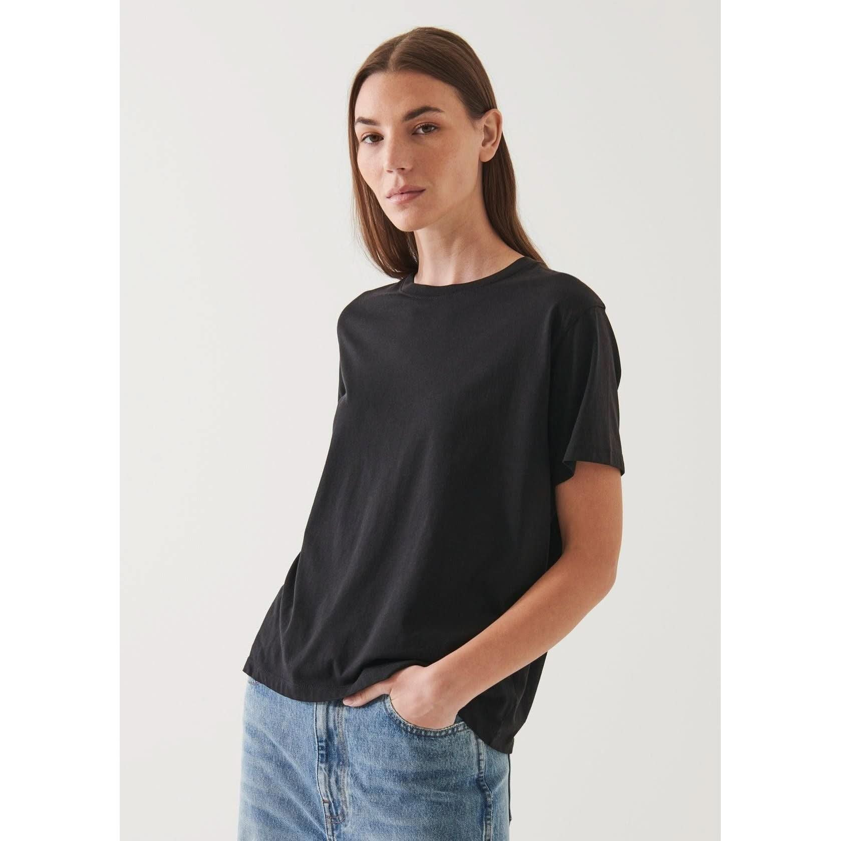 A person with long brown hair, wearing an oversized fit black Patrick Assaraf SS Iconic Boyfriend Crew T-shirt made of soft pima cotton and light blue jeans, stands against a plain light background with one hand in a pocket, looking towards the camera with a neutral expression.