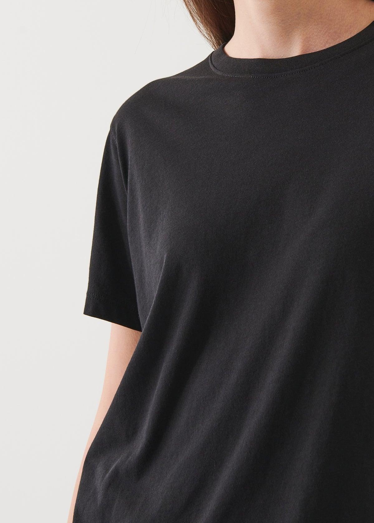 Close-up of a person wearing a plain black Patrick Assaraf SS Iconic Boyfriend Crew. The photo captures the upper torso and part of the left arm, with the head and face partially out of the frame. The background is a neutral light color.