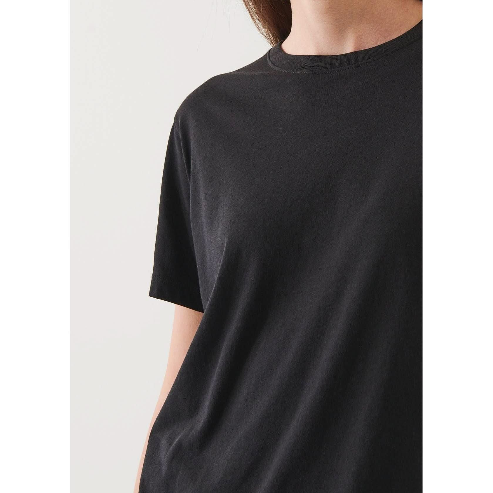 Close-up of a person wearing a plain black Patrick Assaraf SS Iconic Boyfriend Crew. The photo captures the upper torso and part of the left arm, with the head and face partially out of the frame. The background is a neutral light color.