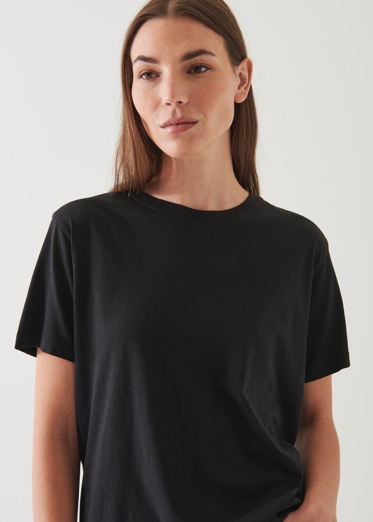 A person with long brown hair and a neutral expression is wearing the Patrick Assaraf SS Iconic Boyfriend Crew Black T-shirt, featuring an oversized fit. They are standing against a light-colored background.