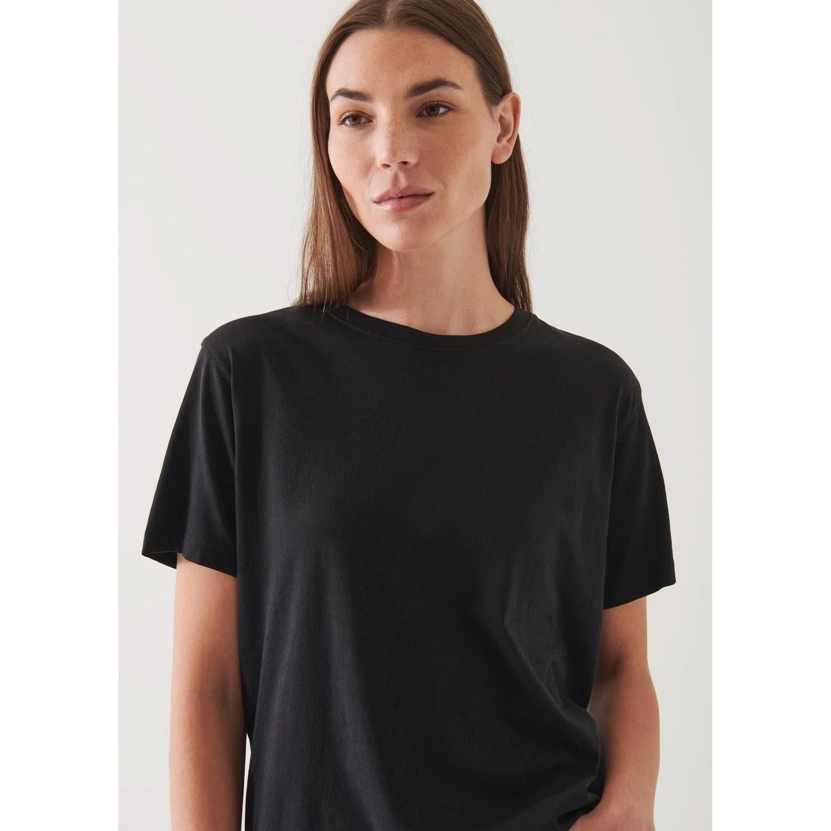 A person with long brown hair and a neutral expression is wearing the Patrick Assaraf SS Iconic Boyfriend Crew Black T-shirt, featuring an oversized fit. They are standing against a light-colored background.