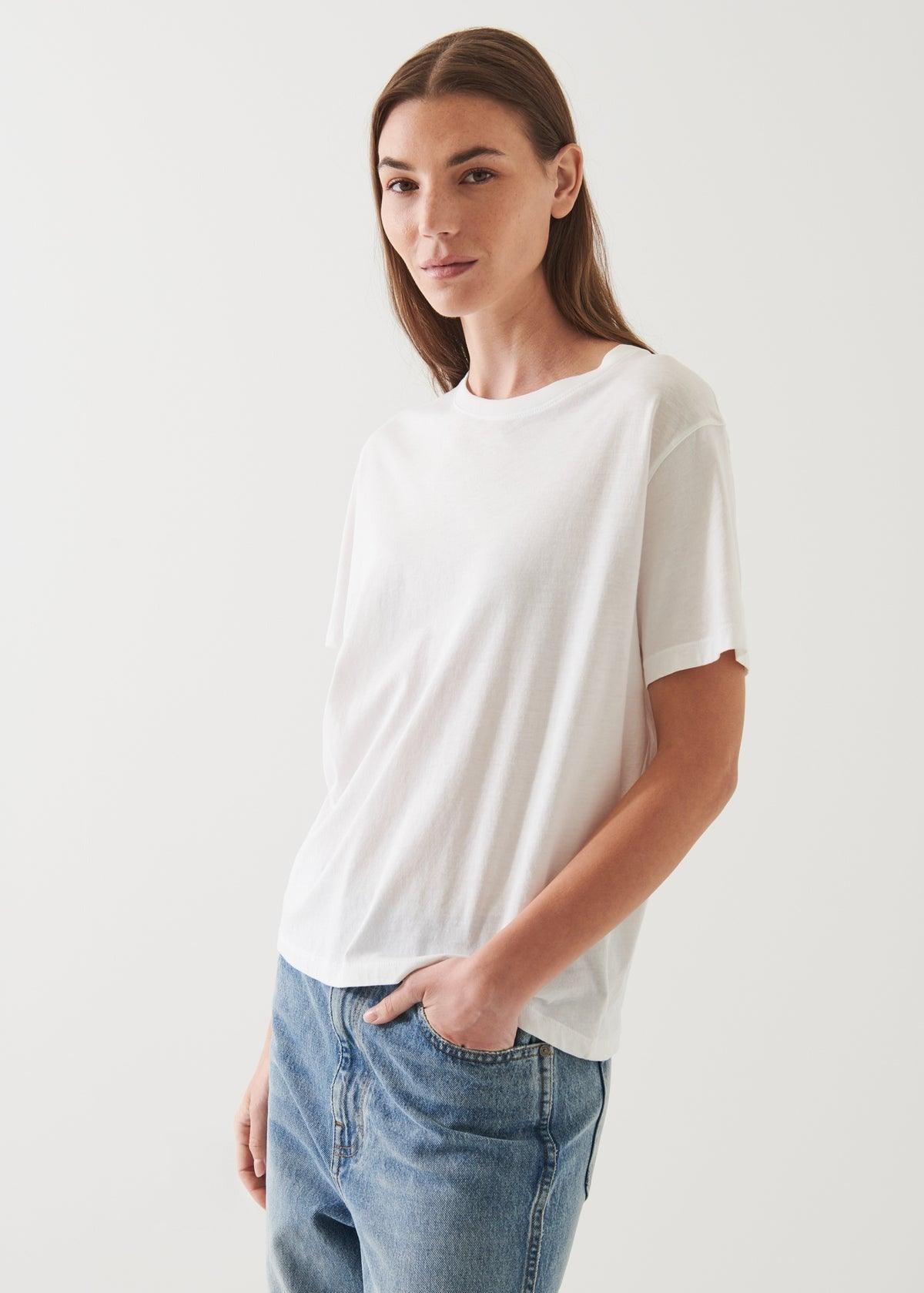 A person with long brown hair is wearing an oversized fit Patrick Assaraf SS Iconic Boyfriend Crew White T-shirt and blue jeans. They are standing against a plain, off-white background, looking towards the camera with a slight smile and one hand in their pocket.