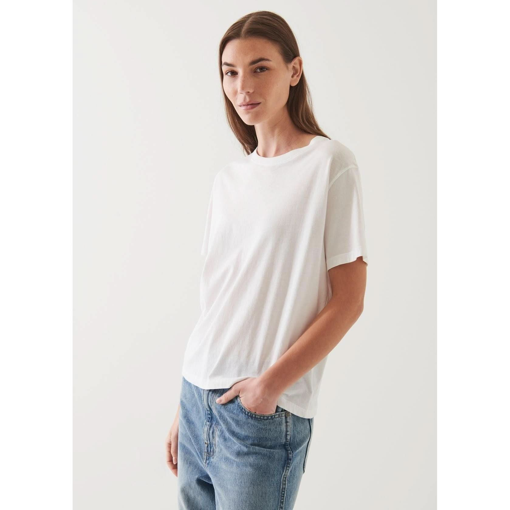 A person with long brown hair is wearing an oversized fit Patrick Assaraf SS Iconic Boyfriend Crew White T-shirt and blue jeans. They are standing against a plain, off-white background, looking towards the camera with a slight smile and one hand in their pocket.