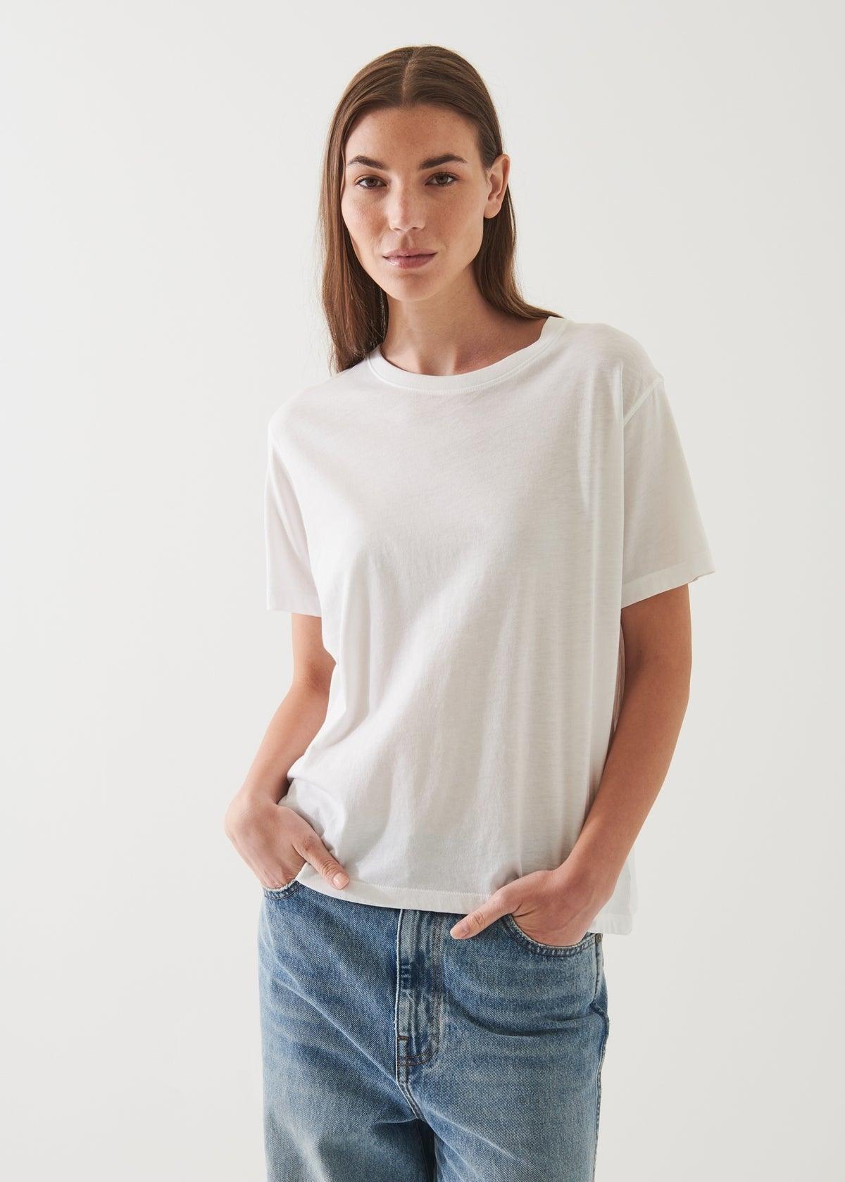 A woman with long brown hair is wearing an oversized Patrick Assaraf SS Iconic Boyfriend Crew White made from soft pima cotton and blue jeans. She is standing with her hands in her pockets, facing the camera against a plain white background.