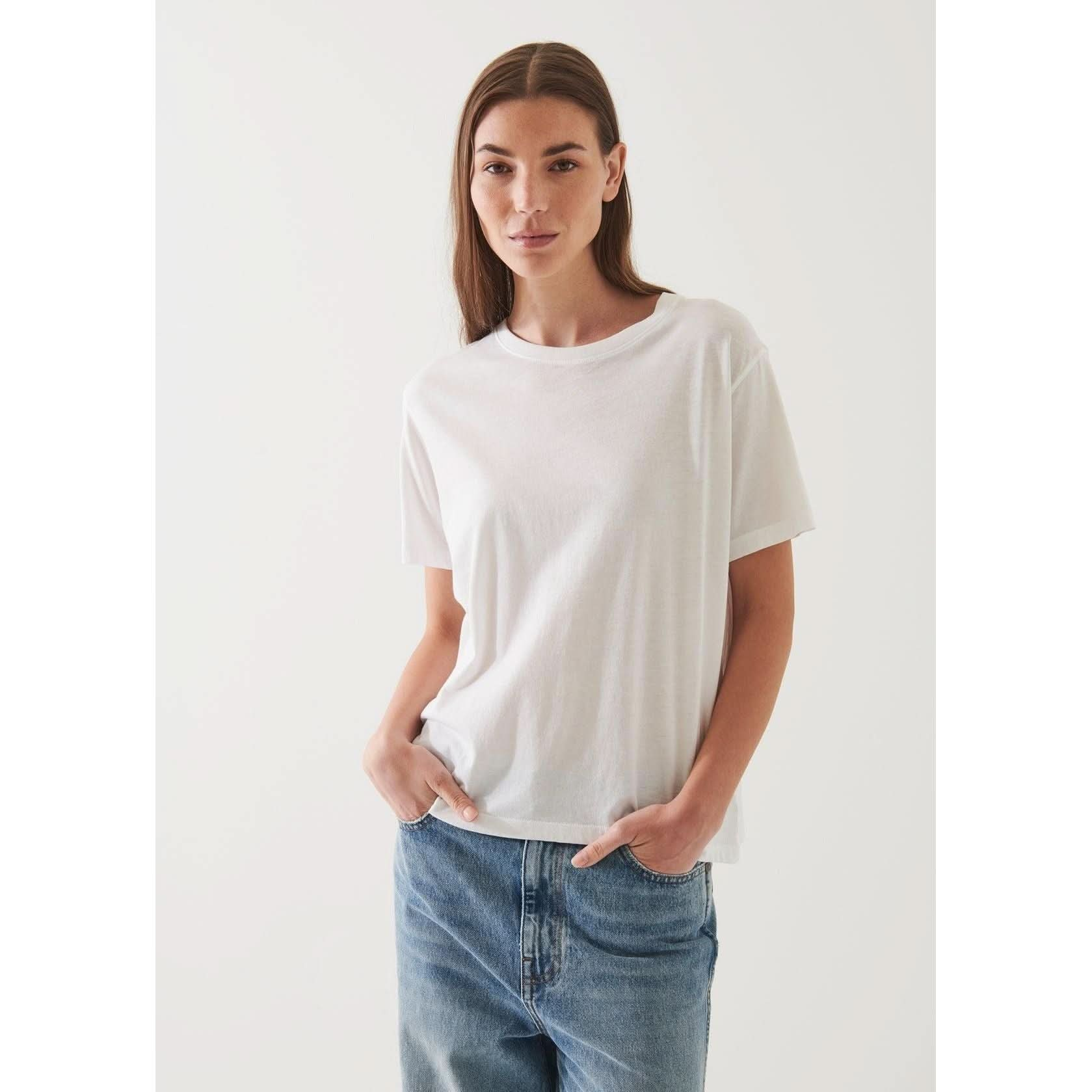 A woman with long brown hair is wearing an oversized Patrick Assaraf SS Iconic Boyfriend Crew White made from soft pima cotton and blue jeans. She is standing with her hands in her pockets, facing the camera against a plain white background.